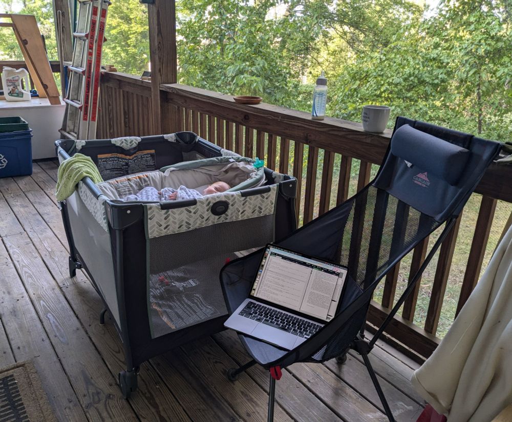A baby sleeps in a bassinet on an outdoor porch. Next to the bassinet is a camping chair with a laptop. Plenty of sunshine and bright green bushes, trees, and grass are visible just outside the porch railing.