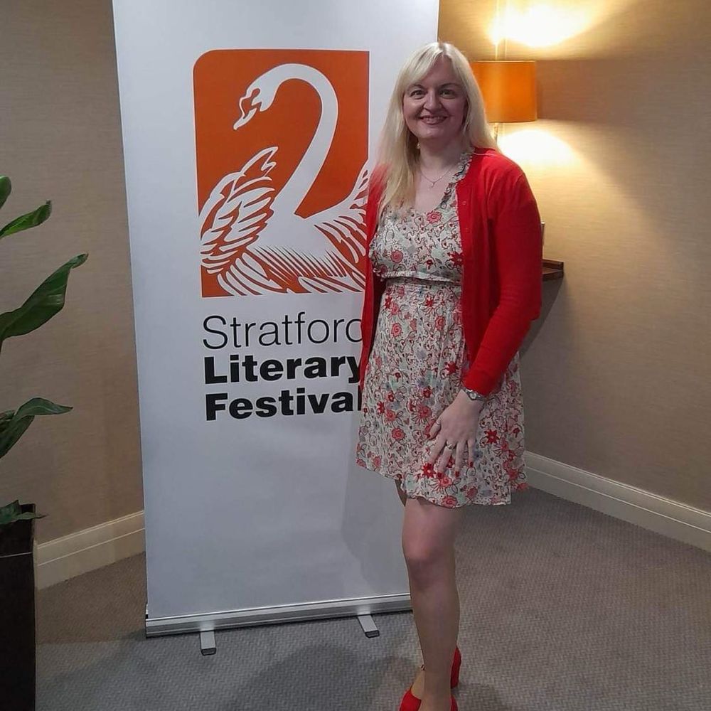 Jo wears a white and red dress and red cardigan and shoes in front of Stratford Literary Festival sign in 2023
