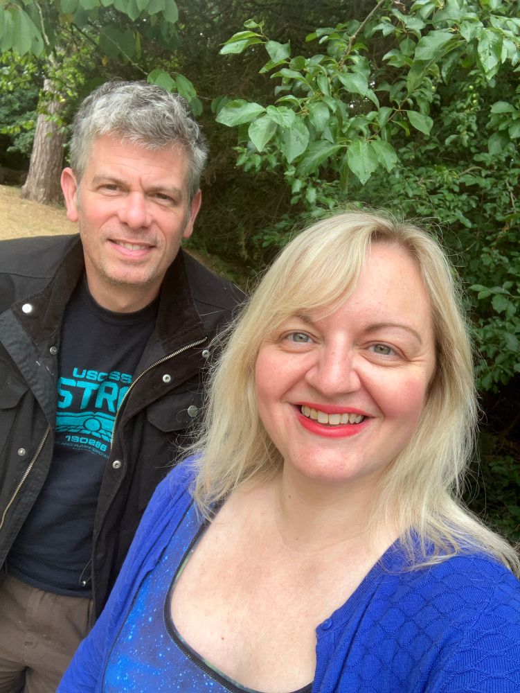 Adam and Jo in a park with a green plant behind them 