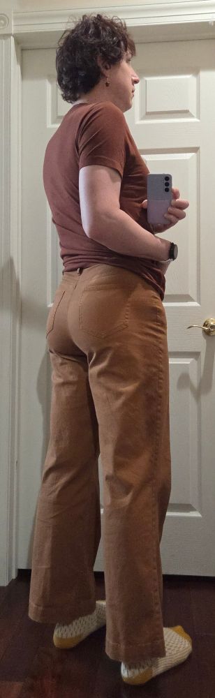 A woman in a brown shirt and tan pants with yellow socks and acorn earrings takes a photo through a mirror of her back.