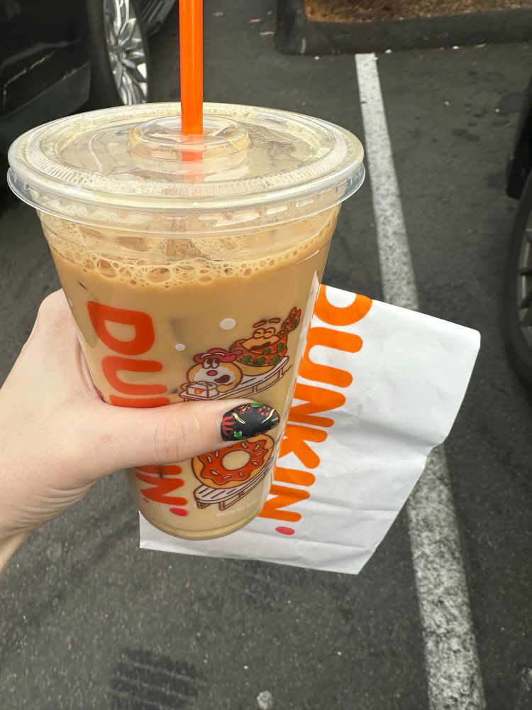 A cup of Dunkin iced coffee and a bag containing a donut being held in a parking lot
