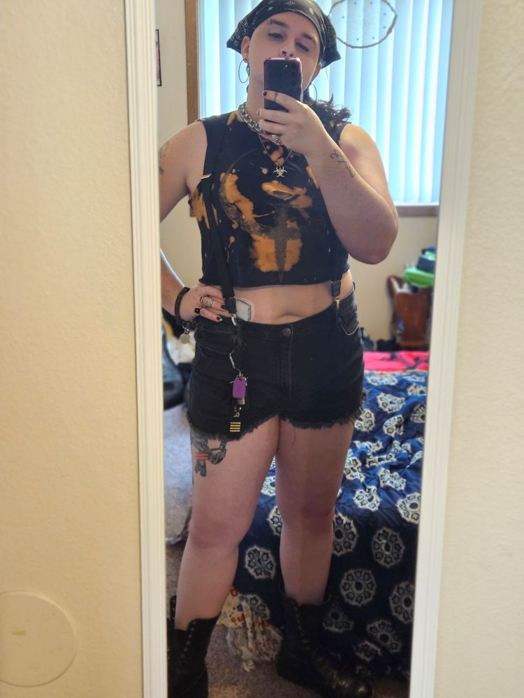 Sabryna taking a mirror selfie. Her hair is in a low side ponytail and she's wearing a black bandana. She has on a typical outfit of a bleached Quake t-shirt, black denim shorts, combat boots, carabiner, and various chains and tarnished silvery jewelry. The twist is she's wearing a pair of black suspenders which extend up from her shorts, resting around her tits before going over her muscular shoulders and behind her back