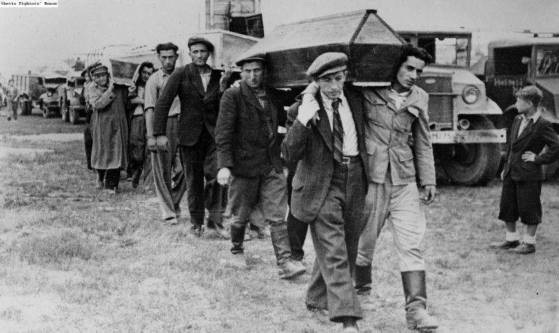 Fotografie zeigt die Beerdigung weniger Tage danach. Männer gehen in einer Reihe und tragen Särge auf den Schultern an großen Fahrzeugen vorbei. Nachweis von wiki commons folgt. 