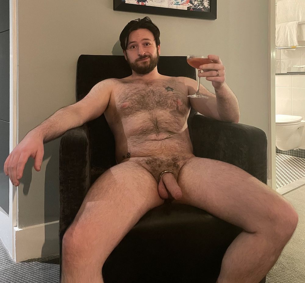 Rossy is leaning back in a chair naked, holding a martini glass 