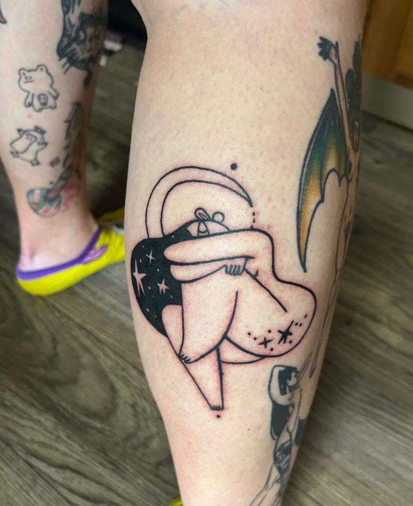 A close-up of Kayleigh’s calf shows a fresh tattoo. It is a black ink illustration of a woman hugging her knee while sitting under a crescent moon. There are stars in her hair and on the back of her thigh. 