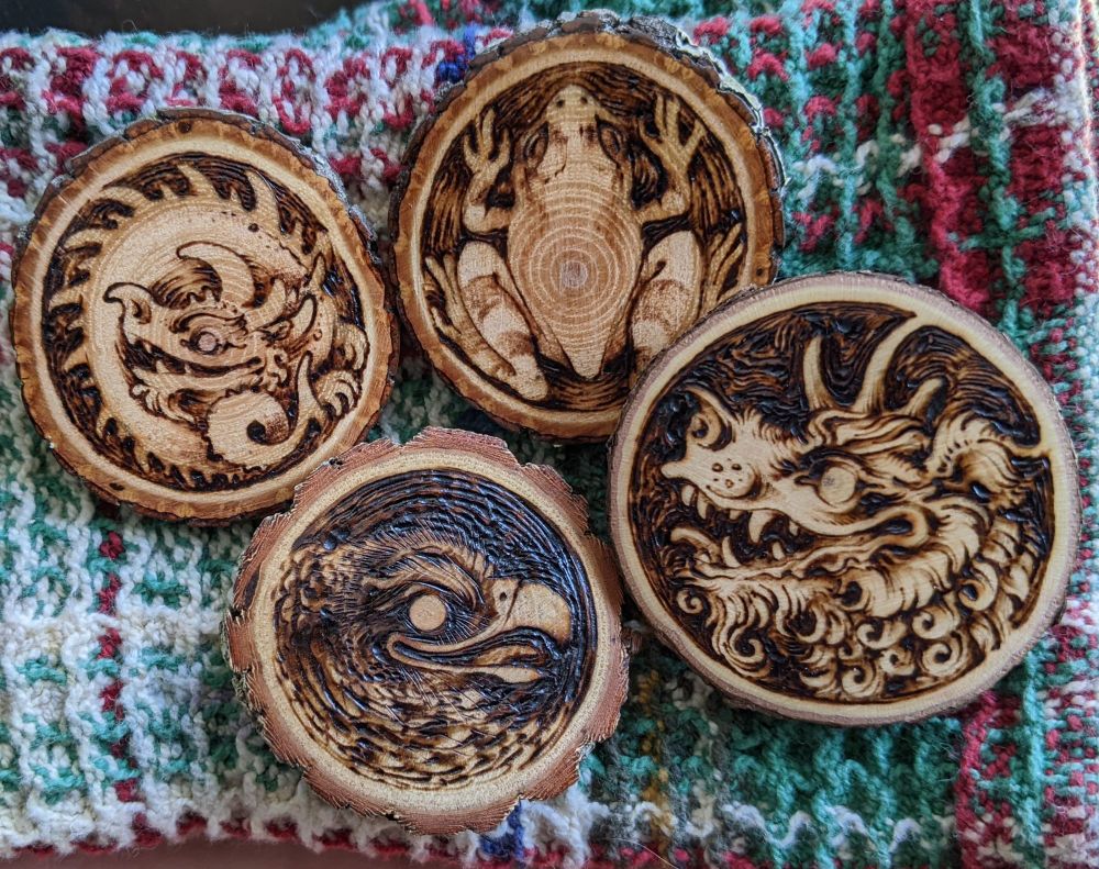 Photo of four slices of wood with pyrography artwork burned on; a dragon, a frog, a bird, and a fuzzy creature 
