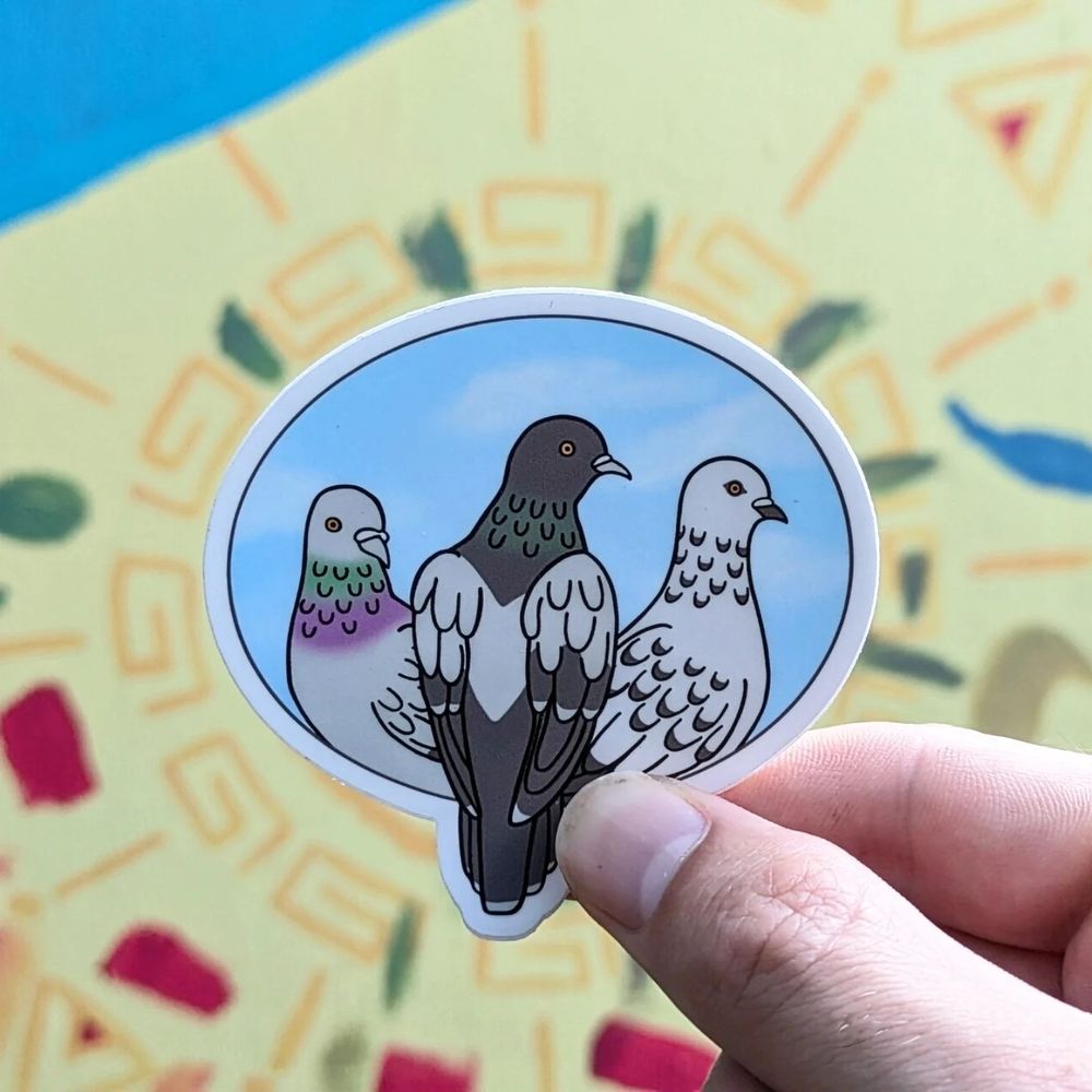 A hand holding a sticker of three pigeons, against a colorful painted backdrop. 