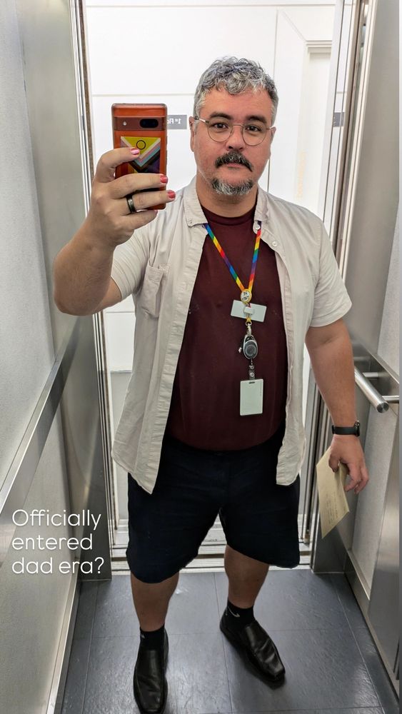 Myself standing inside a lift, taking a selfie. I am wearing black leather shoes, black socks, navy blue shorts, burgundy t-shirt and an opened short sleeved pink shirt on top. I am also wearing glasses and a rainbow harness with my work ID. My right hand is holding an envelope and my left hand is holding my phone. There are words digitally imposed over the picture saying "Officially entered dad era?"