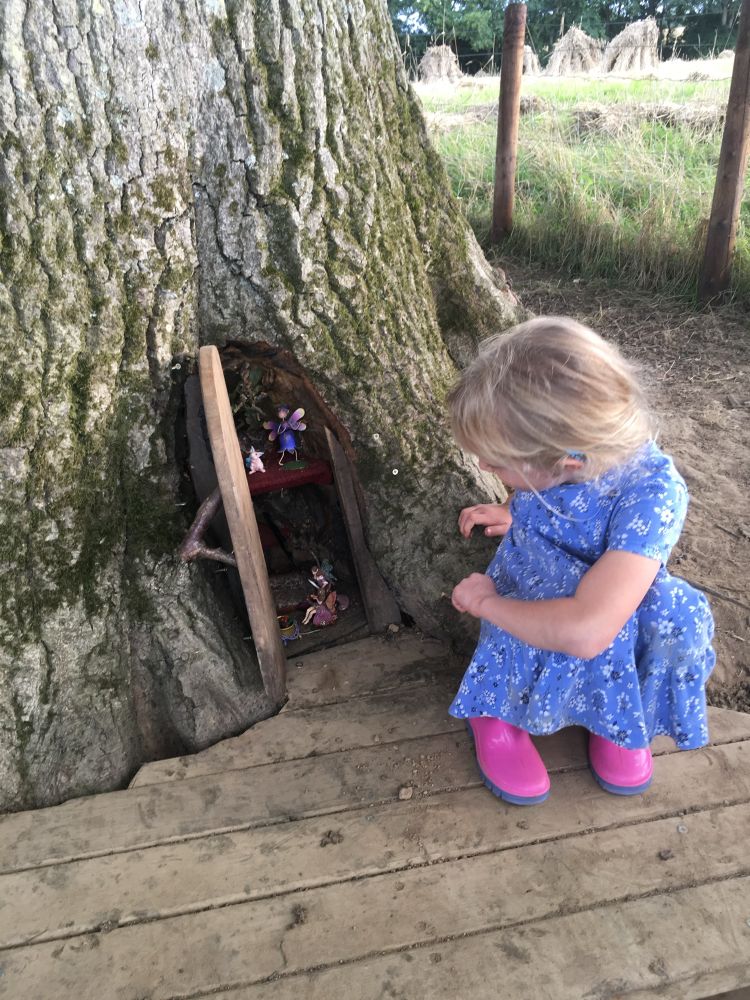 A little girl crouched down at the base of a tree looking into an arched doorway into a fairy’s home. 🧚‍♀️🧚🧚‍♂️