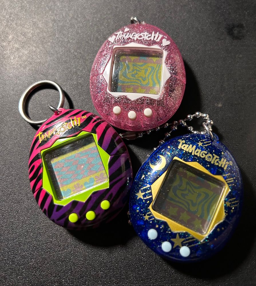 Three Tamagotchi devices, a pink and purple one with green buttons and trim, it has black tiger-like stripes. A pink glitter one with white trim and buttons. And a blue glitter one with gold stars and moons, gold glitter with gold trim, and bluish white buttons.