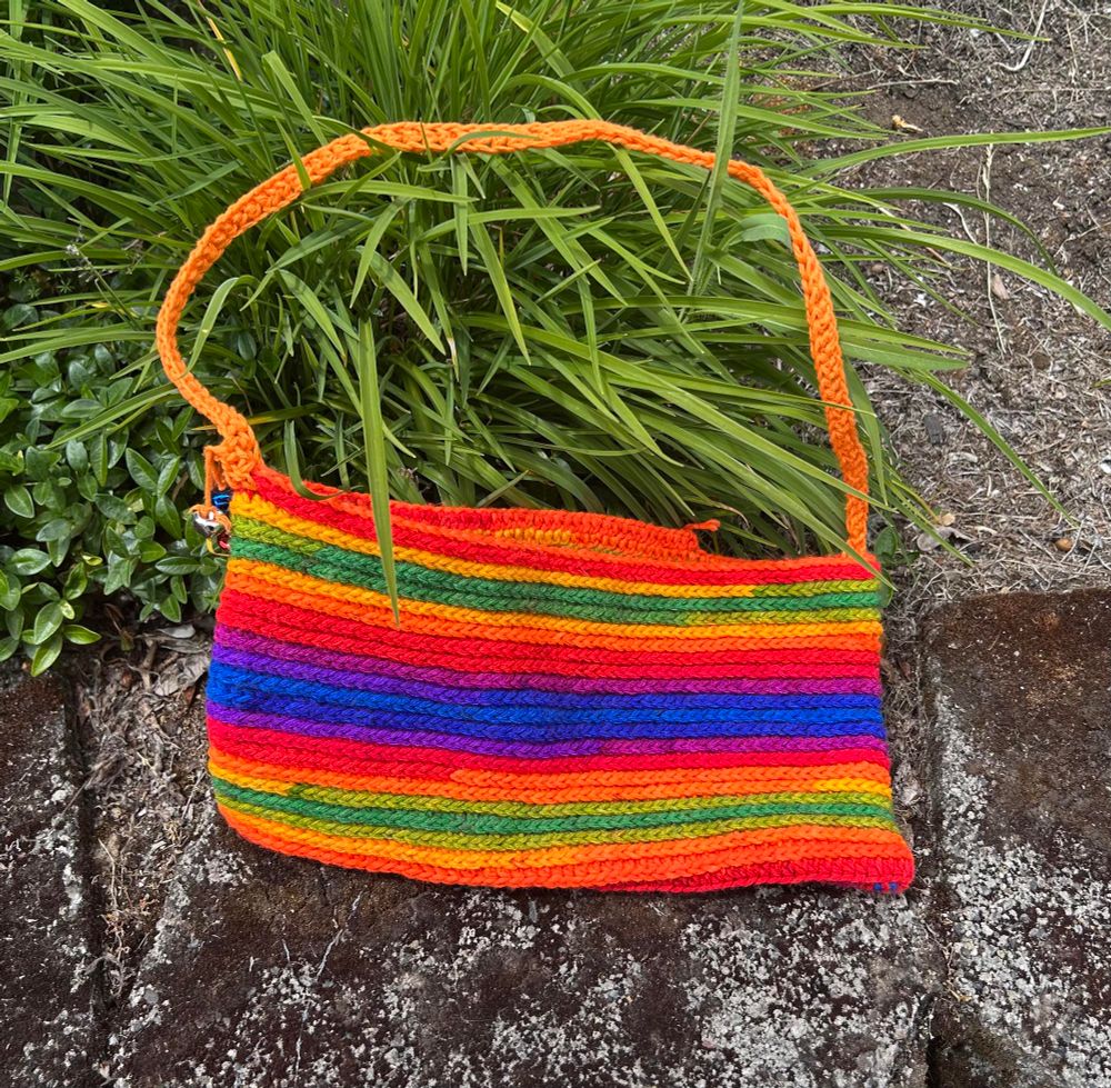 A rainbow crochet bag being propped up by a plant. It had little bells on its left side.
