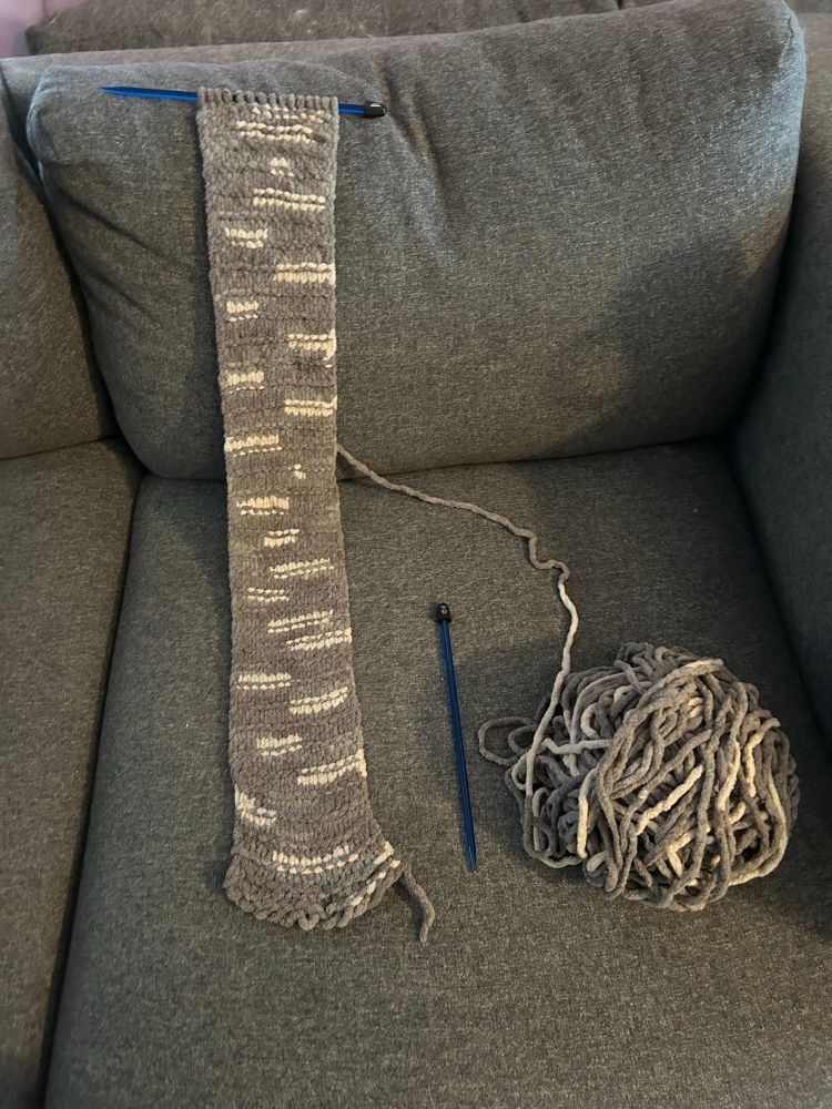 A grey and white scarf being knitted, beside it is a semi messy ball of yarn, and a blue knitting needle. They are resting on a grey couch.