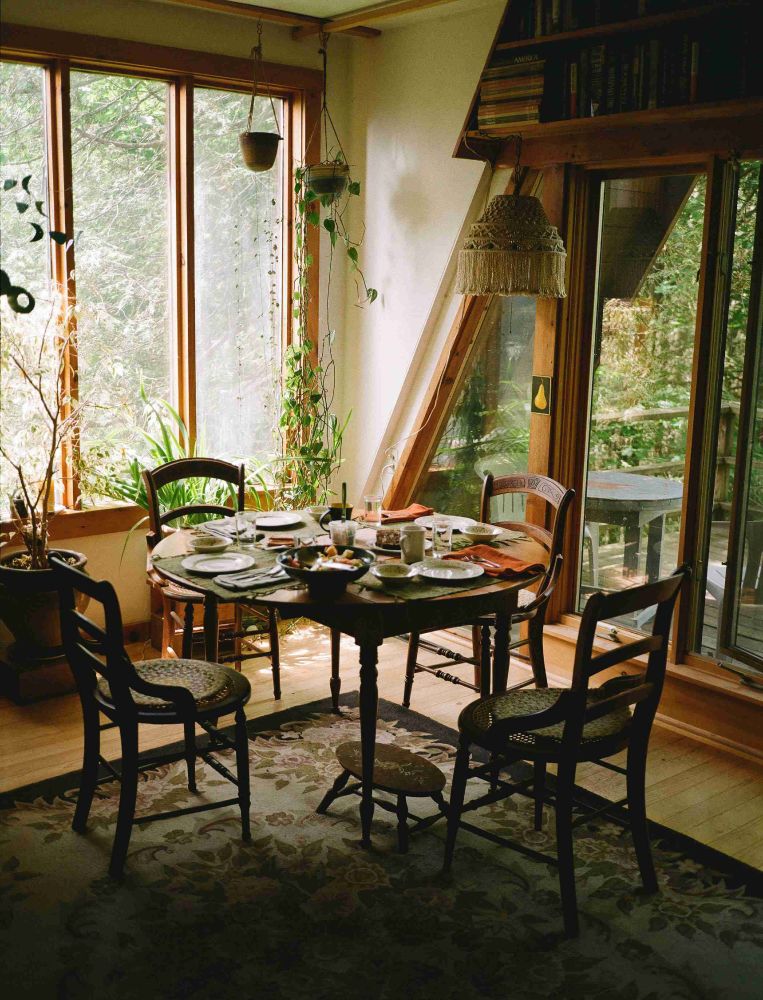 A set table in a dining room, with plants surrounding it and a wall bookshelf behind it. A porch patio is visible in the background.