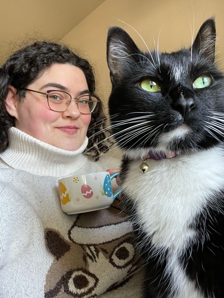 Selfie of Sam Concklin in a turtleneck holding a cup of tea while her male tuxedo cat Saffron sits majestically in the foreground and looks off into the distance