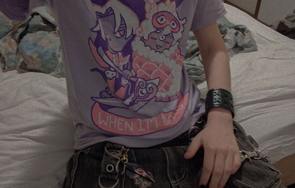 A picture of me sitting down on my bed, the camera is positioned in a way where you can only see my shirt and a small part of my pants. The shirt has two characters from the game Hades on it, Thanatos and Hypnos, and reads "I'll sleep when I'm dead". There are two keychains attached to my pants with the character Blade from Honkai Star Rail on one and Ghostface from Scream on the other, which is shaped like a knife. 