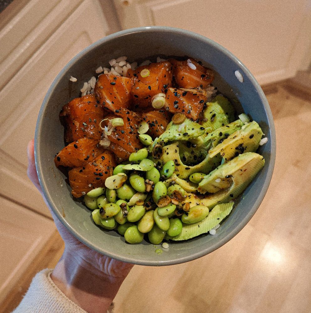 A picture of some food in a bowl shot from above. The contents of the bowl are rice, salmon, avocado, spring onion, and edamame beans with a dressing sauce made of soy sauce, olive oil, sesame seeds, honey, water, and some pepper and salt.