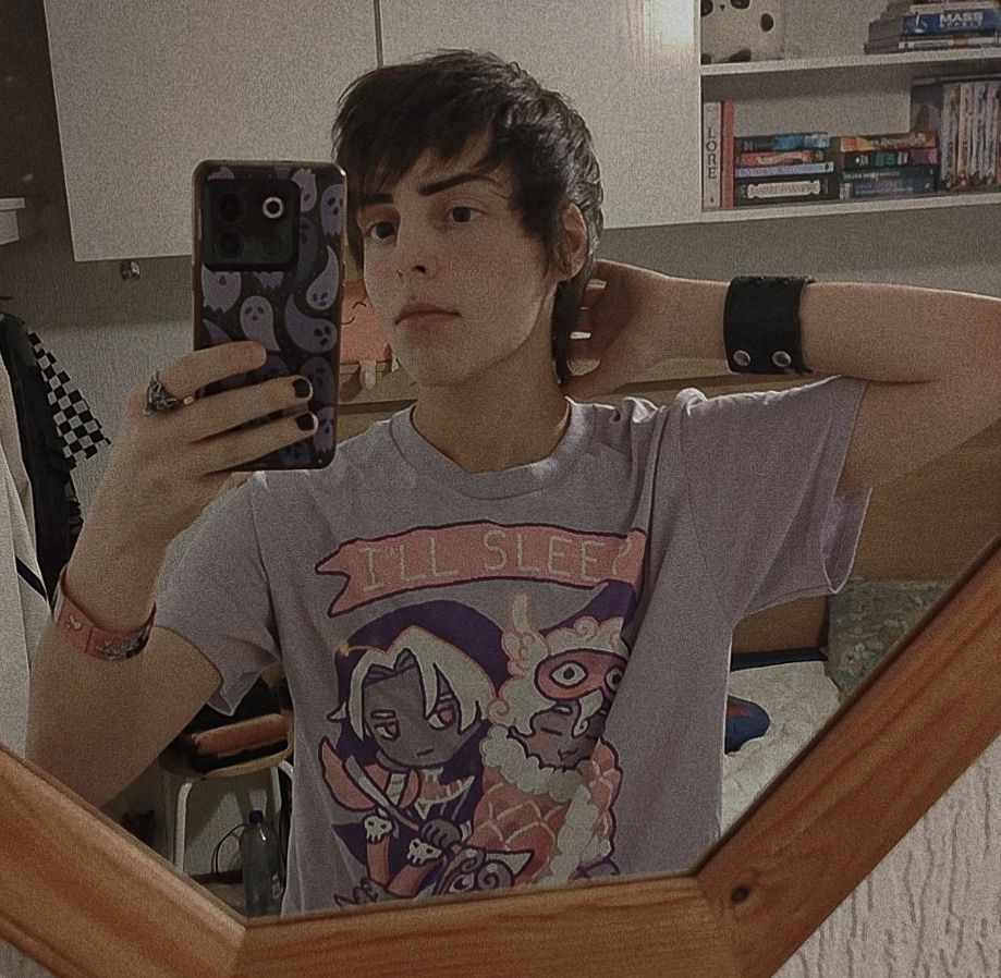 A picture of me looking at the mirror with one of my hands holding up my phone and the other resting behind my neck. I'm wearing a pink shirt with two characters from the game Hades, Thanatos and Hypnos. It reads "I'll sleep when I'm dead" 