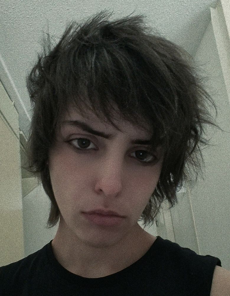 A picture of me staring into the camera. I'm frowning a bit, my hair is cut into a messy emo style with bangs. I'm wearing dark eyeliner on the bottom of my eye. 