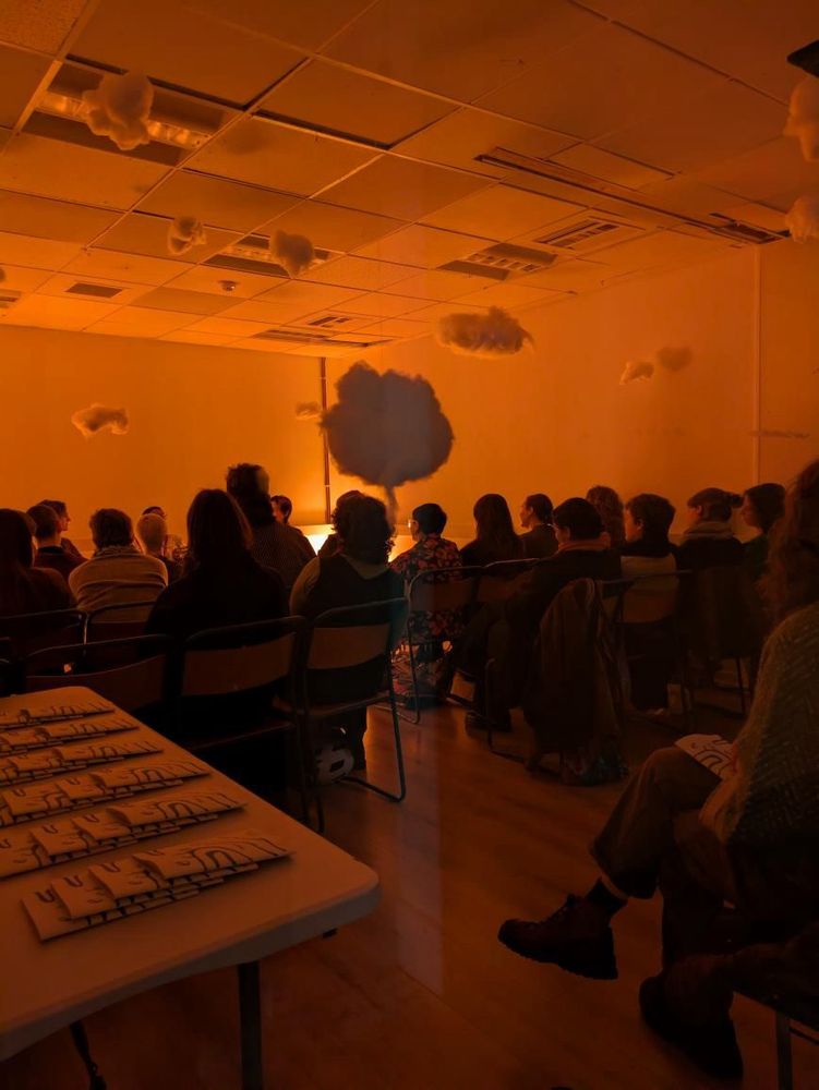A photo of people sitting in a room basked in a warm orange glow, with cotton clouds hanging from the ceiling. There are zines on a table to the side.
