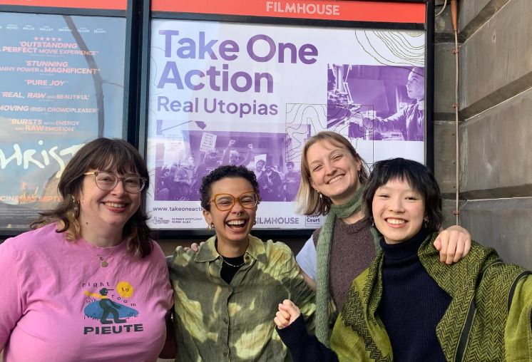 A group of four smiling and laughing people standing in front of a Take One Action Real Utopias poster