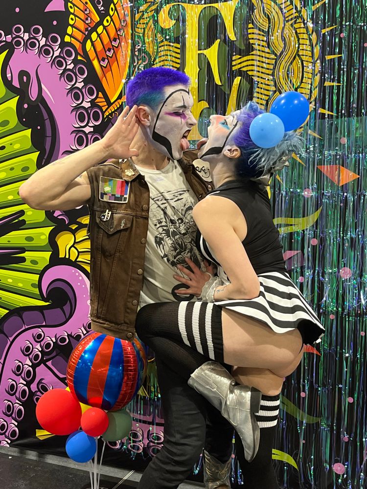 Two punk rock clowns express love by sticking out their tongues and showing the other the back of their throat 