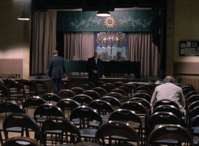 ChatGPT describes the image as "The image depicts a small, sparsely populated auditorium or community hall with rows of empty chairs. The stage is centrally positioned, decorated with a colorful backdrop featuring a large painted scene and a decorative sun-shaped ornament hanging above. Two men in suits are on the stage, engaged in conversation or preparing for an event, while a third man sits alone in the audience, facing away from the stage. The setting has a somewhat somber or reflective atmosphere, with subdued lighting and a simple, functional design suggesting a local gathering or meeting. The overall tone hints at a moment of anticipation or introspection within a community context."

BotOfDarkness, version 2.0.23. AWS Lambda version: 1.14.