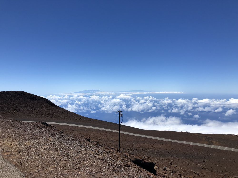 Photograph I took from the summit of Haleakalā in Haleakalā National Park. Brown foreground of lava rock and the clouds and ocean far below. The peak sits at 10,023 ft (3,055 m) above sea level.