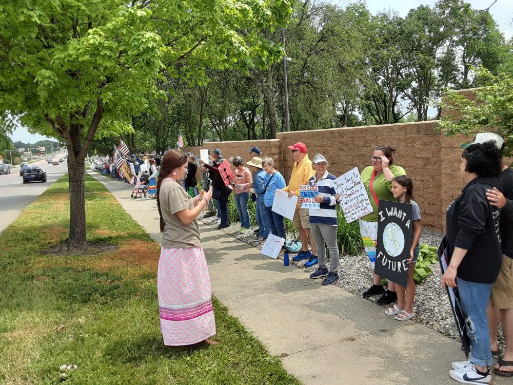 A group of local citizens stand with signs, lining the sidewalk in front of a wall. 