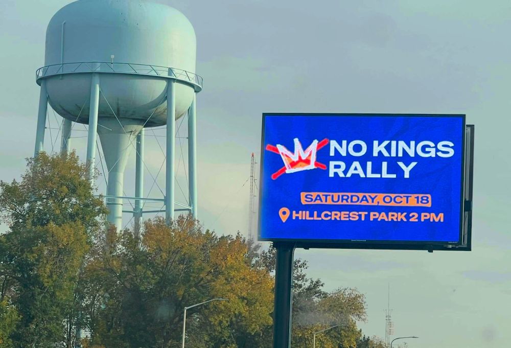 A picture of a roadside display near a water tower. The sign reads, "No Kings Rally Saturday, Oct 18th Hillcrest Park 2pm". 