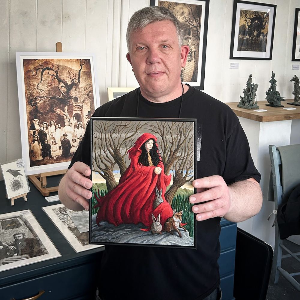 Photograph of Welsh artist David W. J. Lloyd holding up his painting Melangell's Cloak in a gallery filled with his work