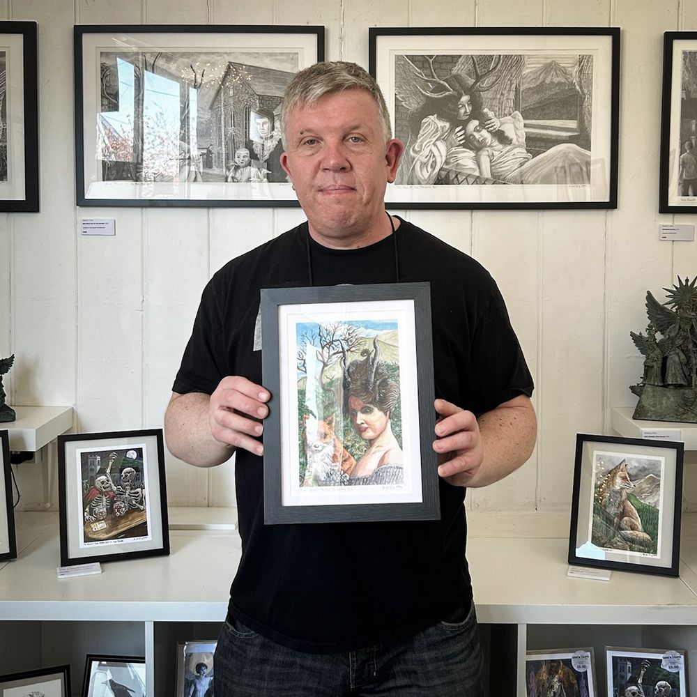 Photograph of Welsh artist David W. J. Lloyd holding up his painting The Queen Of The Tylwyth Teg in a gallery filled with his work