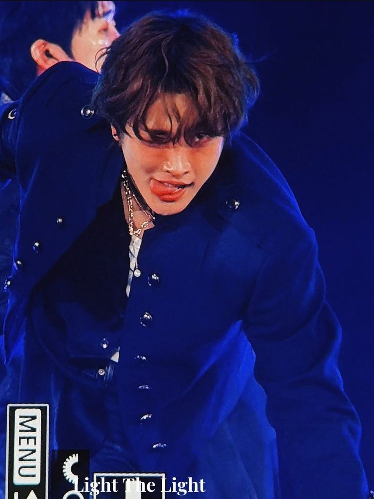 A photo of hongjoong on stage. He's wearing the same blue coat as in the first photo in this post and the same hairstyle as in the other photos. He's leaning forward and looking straight ahead. He's smiling and winking and his tongue is poking out of the side of his mouth. 