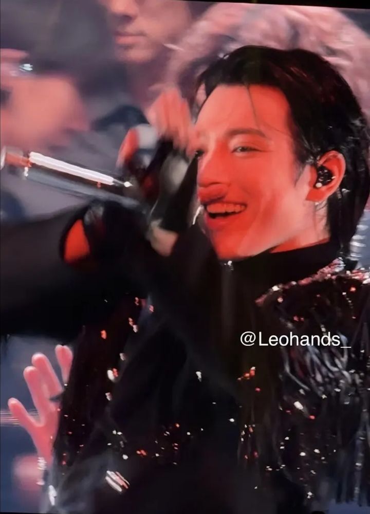 A photo of Hongjoong on stage. He is wearing a sparkly black outfit and holding up the mic to his mouth while waving with his other hand. His hair is black and combed back and his signature beautiful smile is on his face. 