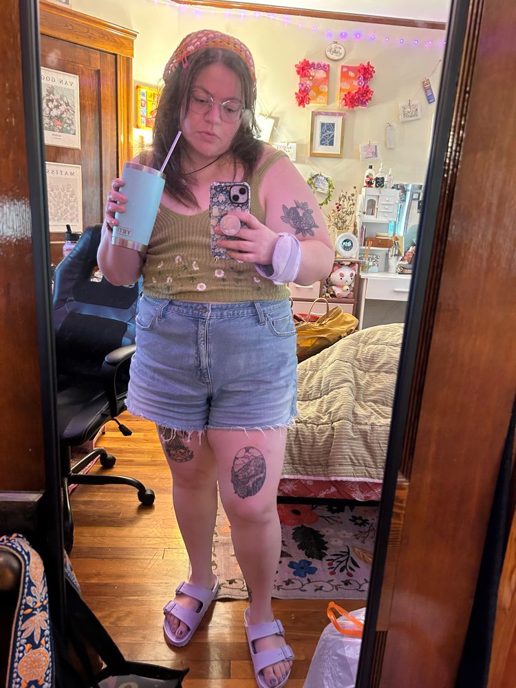 mirror selfie holding my iced coffee. i’m wearing high-waisted light denim shorts, purple house sandals, a green knit tank, a crocheted pink, orange and white bandana, and a purple ice pack on my wrist. it’s chronic pain but in a sexy summertime way