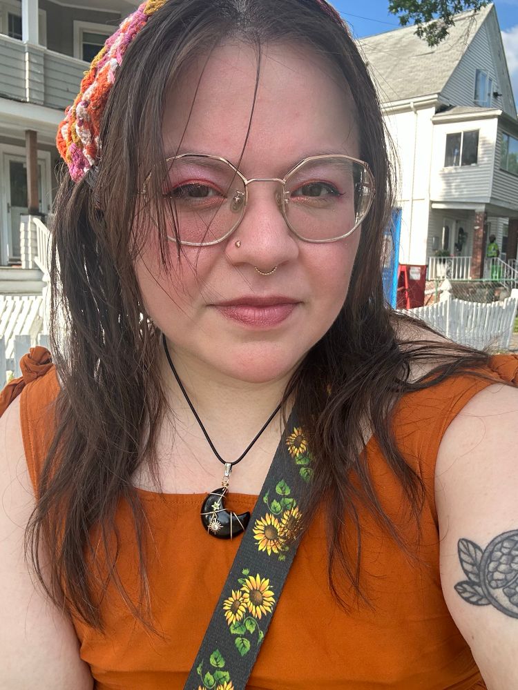 i’m wearing an orange sleeveless dress with a sunflower bag strap across my chest, a crocheted pink, orange and white bandana, and a black moon pendant necklace 