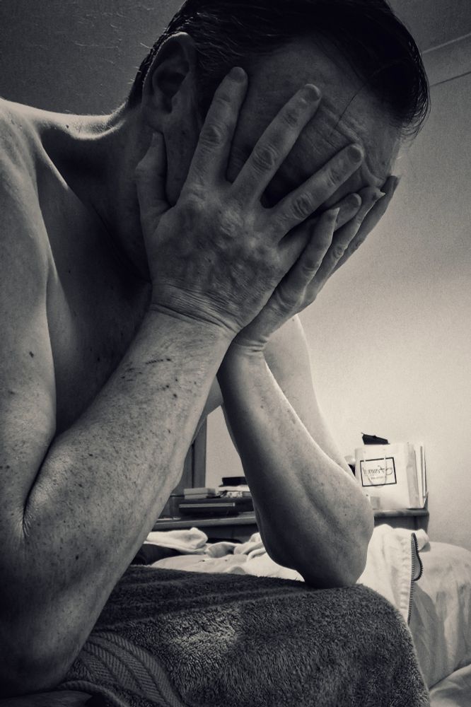 Mono pic of naked white male sitting on the edge of a bed, head in hands, close cropped because this week is going to fuck him over.