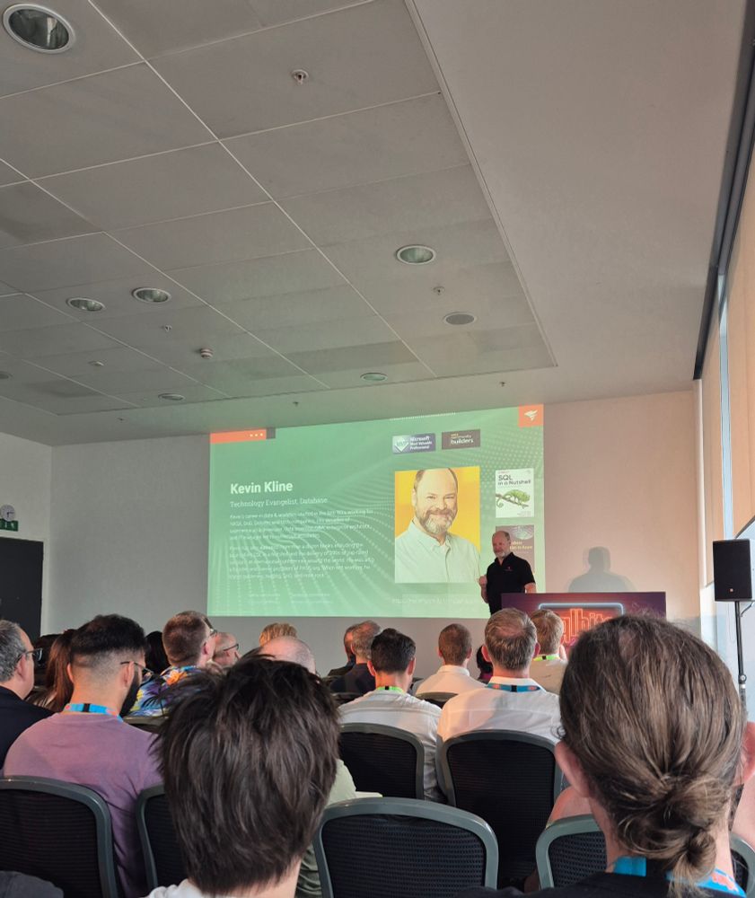 Rows of people in seats before a presenter in front of his slides, with a intro slide on the screen