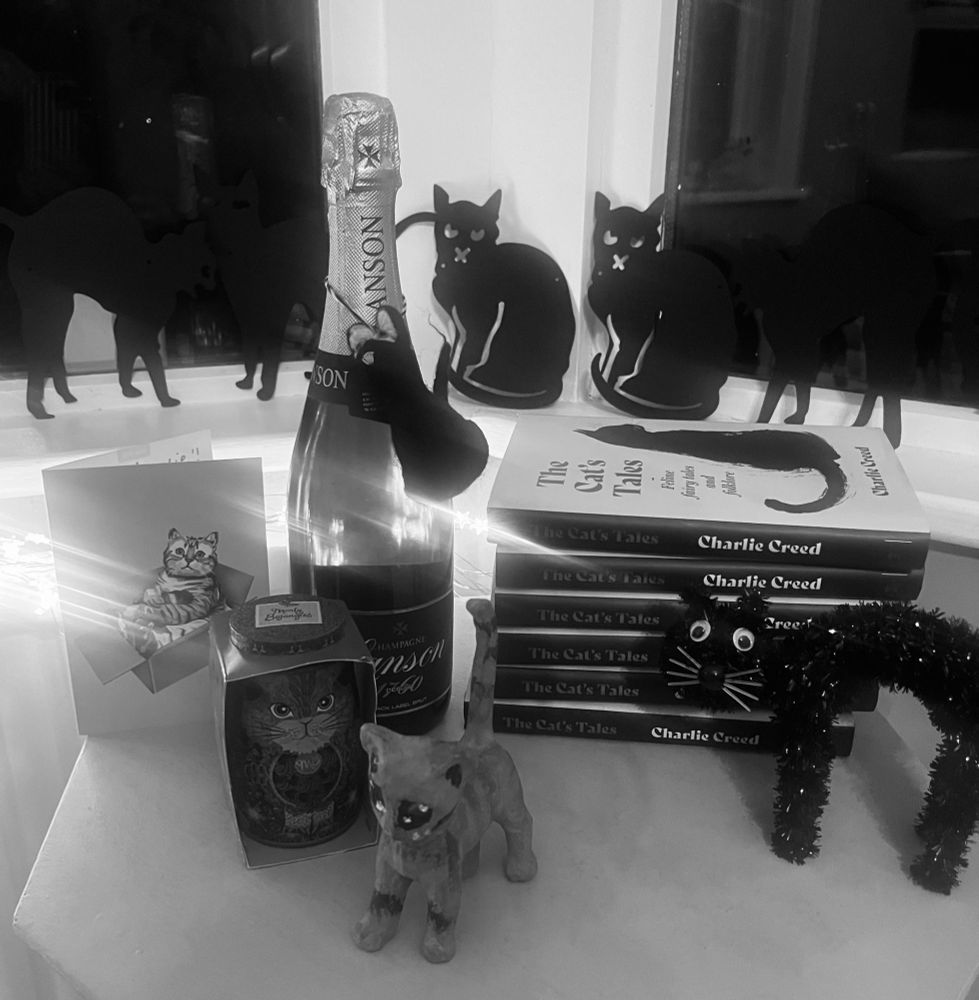 A pile of The Cat's Tales: Feline Fairy Tales and Folklore surrounded by toy cats, a card, and a bottle of champagne