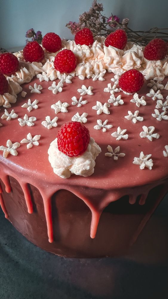 An angled top-down view of a chocolate ganache-covered cake with ruby ganache top & drips down the sides. The top of the cake is decorated with small whipped cream flowers & swirls, and fresh raspberries. 