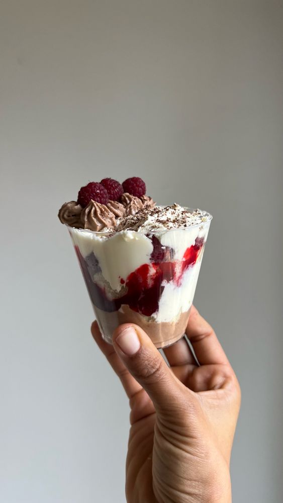A hand holding up a trifle of chocolate mascarpone mousse, white cake, raspberry jam, fresh raspberries, and whipped cream against a plain white background. 