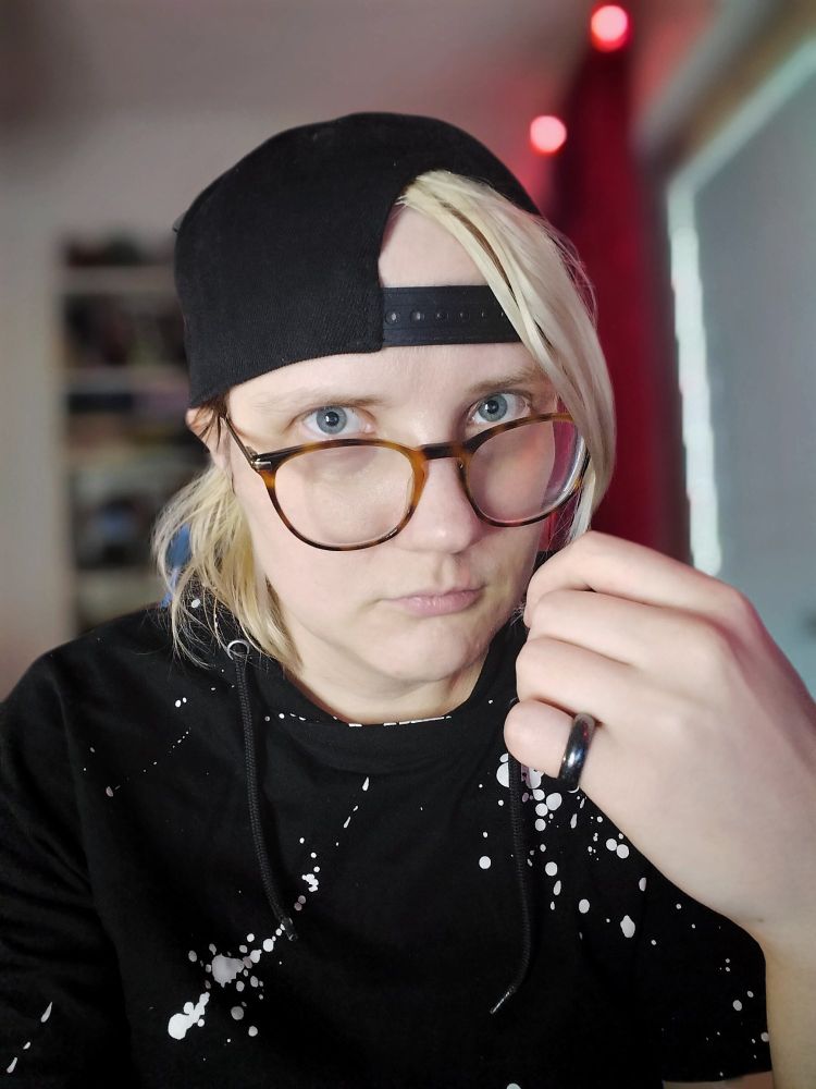 A blonde person, wearing black backwards cap, black t shirt with white splatter print.