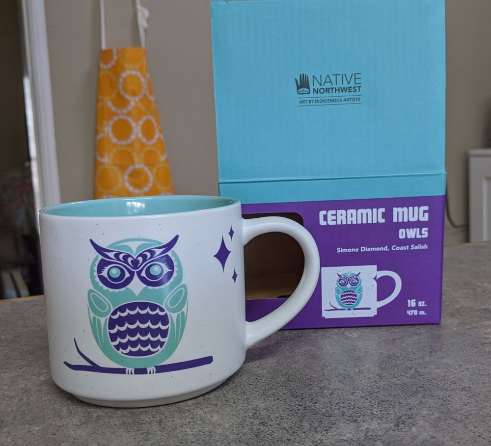 Picture of a beautiful 16oz white mug with a Coast Salish style teal and purple owl on it. The interior of the mug is the teal colour.

Beside the mug is a box. It labels the mug as coming from the Native Northwest company and the artist is Simone Diamond of the Coast Salish.