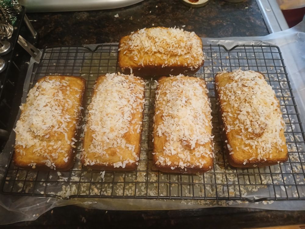 Five loaves of golden-brown piña colada bread, glazed and topped with toasted coconut, are cooling on a rack.