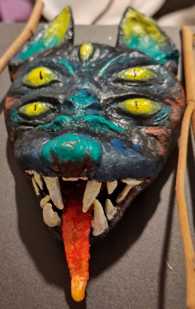 a clay sculpture of the face of a doglike monster with big sharp teeth and five eyes. black with teal accents, eyes and eartips are yellow-green, tongue is a red to orange gradient.