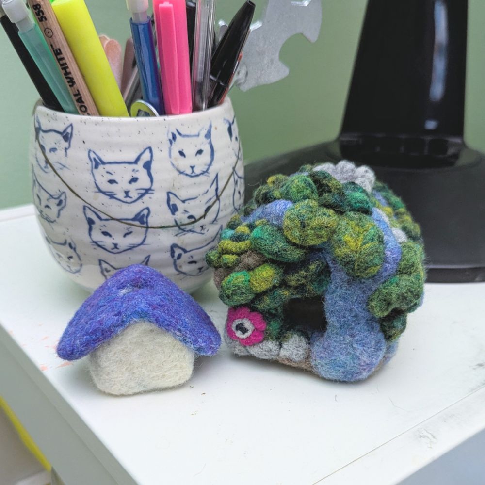 a very tiny needle felted house with a purple roof and blank beige walls next to a slightly bigger but still quite small needle felted house that's been decorated to look like it's being consumed by nature. Behind them is a coffee mug holding various pens and pencils and the base of a fan. 