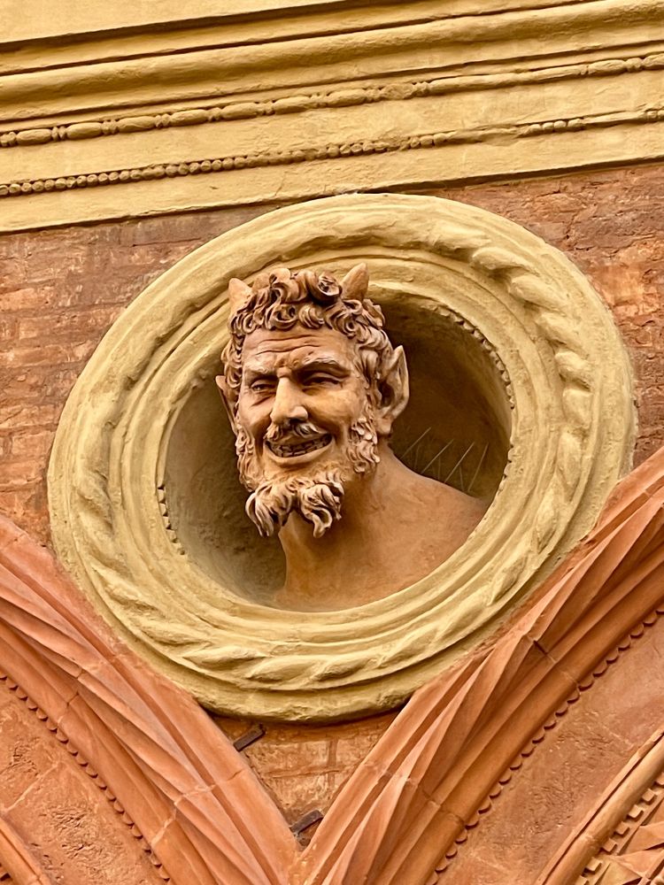 Devil like head sculpture protruding out of decorative circle border on the side of a building.