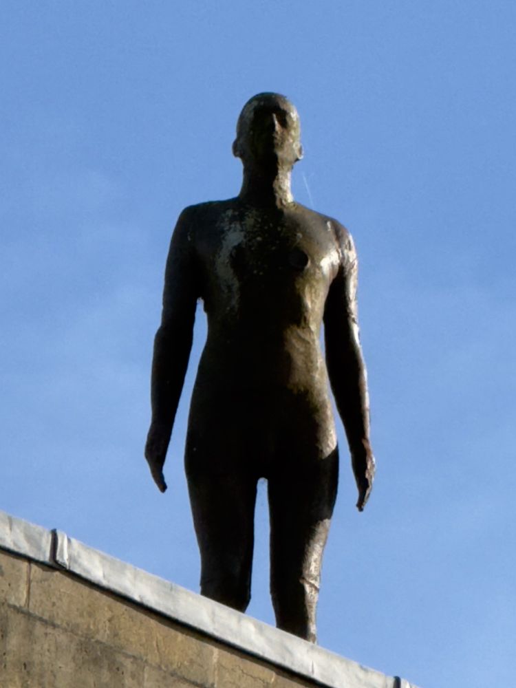 Weathered bronze sculpture of a naked man, stood bolt upright on top of a building ominously looking down. 