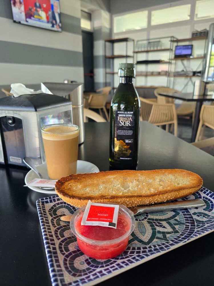 Coffee is a glass next to a plate with a baguette cut in half, toasted, a container filled with grated tomatoes, a pack of salt, and a bottle of olive oil. 