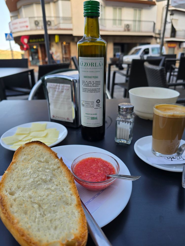 A toasted slice of bread with a small bowl with grated tomatoes, a plate of cheese in the back, a bottle of olive oil, and salt shaker and coffee in a glass. 
