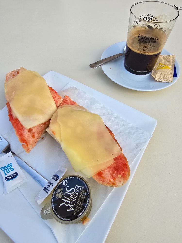 Slices of bread topped with tomatoes, and melted cheese on top, next to a container with olive oil. Behind there is a black coffee. 